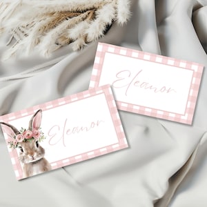 May include: Two place cards with a pink gingham border and the name "Eleanor" in cursive. One card features a watercolor illustration of a bunny wearing a floral crown. The cards are on a white satin surface.