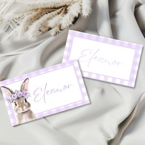 May include: Two place cards with a lavender gingham border and the name "Eleanor" in cursive. One card features a watercolor illustration of a bunny wearing a floral crown. The cards are on a white satin surface.