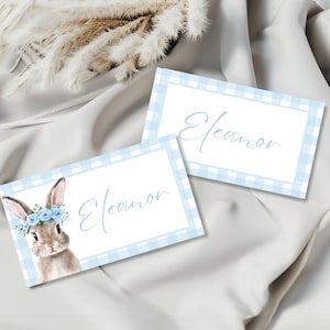 May include: Two light blue and white gingham place cards with the name "Eleanor" in a delicate script. One card features a watercolor illustration of a bunny wearing a floral crown. The cards are on a white satin surface.