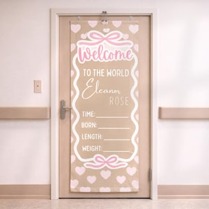 Op de afbeelding: Een beige deurvlag met roze details, met de tekst "Welcome to the World Eleanor Rose". De vlag heeft roze strikken en hartjes. Er zijn vakken om de tijd, geboortedatum, lengte en gewicht van de baby in te vullen.