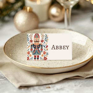 May include: A place card with the name "Abbey" on a cream background, featuring a colorful nutcracker illustration. The card is set on a speckled plate with a gold rim, atop a linen napkin, with festive decorations.