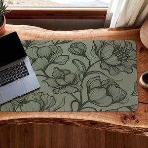 May include: A floral patterned desk mat featuring a detailed botanical design in shades of green and gray. The mat is rectangular and has a laptop on the left side. The design includes large flowers and leaves.