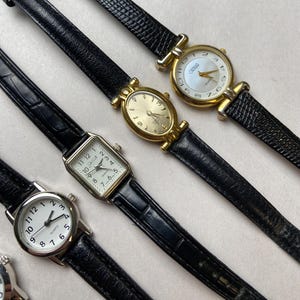 May include: Several vintage wristwatches with black leather bands and gold or silver-toned cases. The watch faces are white or gold, with Roman or Arabic numerals. Some watches have a rectangular or oval shape.