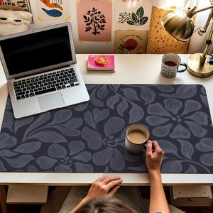 May include: A desk setup featuring a laptop, coffee mug, and a floral patterned desk mat. The mat is dark gray with a repeating flower and leaf design. A gold desk lamp and potted plant are also visible.