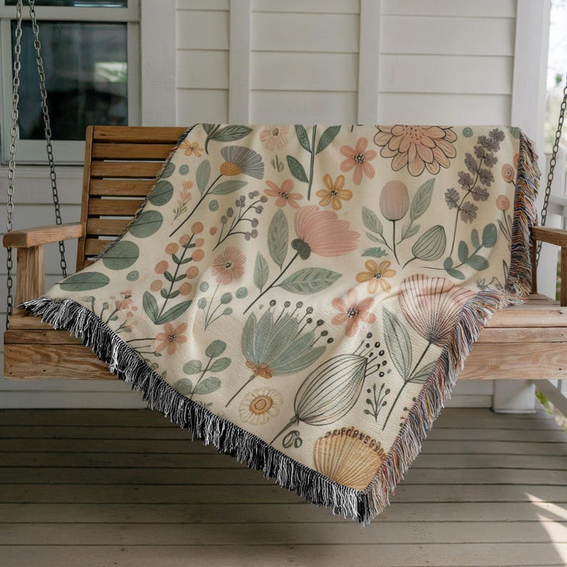 May include: A cream-colored woven throw blanket with a floral pattern featuring pink, yellow, and green flowers. The blanket has a fringe border and is draped over a wooden porch swing.