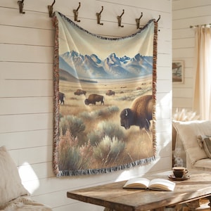 May include: A woven tapestry wall hanging featuring a scenic landscape with a herd of bison grazing in a field with mountains in the background. The tapestry has a fringed border.