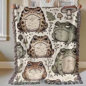 May include: A woven throw blanket with a white background and a repeating pattern of brown and green toads with black eyes. The toads are surrounded by small brown mushrooms, green leaves, and white flowers.