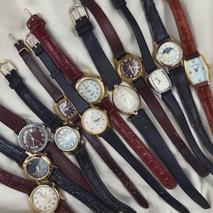 May include: A collection of vintage wristwatches with various band colors, including black, burgundy, and brown. The watch faces feature different designs, with gold and silver tones. Some watches have leather straps, while others have metal clasps.