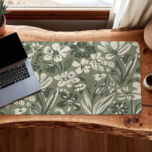 May include: A floral patterned desk mat featuring a design of white and dark green flowers and leaves on a muted green background. A laptop sits on the mat, which is placed on a wooden desk.