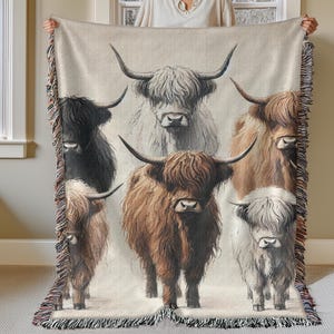 May include: A woven tapestry throw blanket featuring a colorful illustration of six Highland cattle with long, shaggy fur. The cattle are arranged in a row, with their heads facing forward. The background is a light beige color.