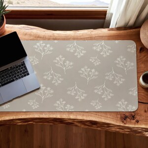 May include: A rectangular desk mat with a beige background and a repeating white floral pattern. A laptop sits on the mat, which is placed on a wooden desk. The desk is near a window with a view of mountains.