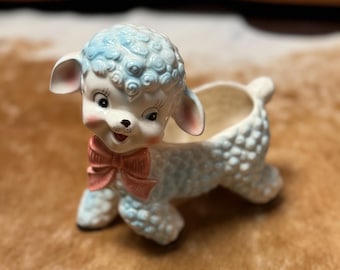May include: A ceramic planter shaped like a white lamb with blue curly fur. The lamb has a pink bow and a happy expression. The planter has a small opening on the top for planting.