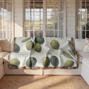 May include: A white throw blanket with a woven design of green olives and olive branches. The blanket has a fringe border.