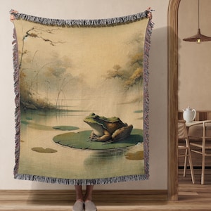 May include: A woven tapestry throw blanket featuring a green frog sitting on a lily pad in a pond. The background is a watercolour-style painting of a misty forest.