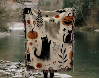 May include: A woven throw blanket with a Halloween theme. The blanket features a black cat, a ghost, pumpkins, and other spooky details. The blanket is beige with orange, black, and white accents.
