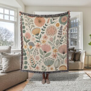 May include: A woven throw blanket with a floral pattern featuring pink, yellow, and green flowers on a cream background. The blanket has a fringe border.