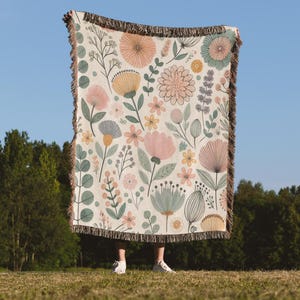 May include: A woven throw blanket with a floral pattern featuring pink, yellow, and green flowers on a cream background. The blanket has a fringe border.