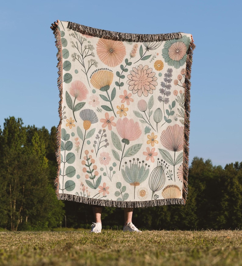May include: A woven throw blanket with a floral pattern featuring pink, yellow, and green flowers on a cream background. The blanket has a fringe border.