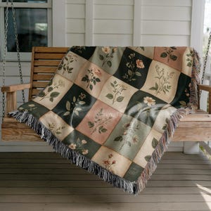 May include: A floral throw blanket draped over a wooden swing. The blanket features a patchwork design with botanical illustrations in shades of green, pink, and cream. The edges are finished with a fringe.