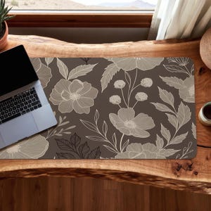 May include: A rectangular desk mat with a floral pattern. The mat features a dark brown background with a repeating design of large, light beige flowers and leaves. A laptop and a small cup are on the wooden desk.