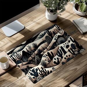 May include: A black and white illustrated mousepad featuring a desert landscape with cacti and other desert plants. The background is a light beige color.