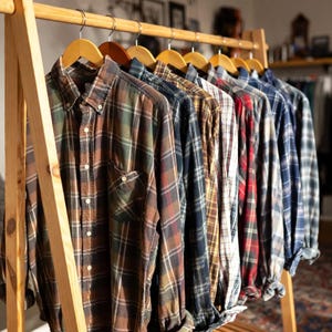 Peut inclure: Une rangée de chemises en flanelle à manches longues et boutonnées, présentant divers motifs à carreaux et couleurs, dont le marron, le vert, le bleu, le rouge et le blanc, suspendues sur des cintres en bois sur un portant en bois clair.