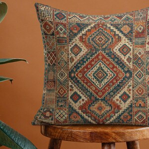 Può includere: Cuscino quadrato con un motivo geometrico nei toni del rosso, blu, arancione e beige. Il cuscino è posto su uno sgabello di legno, mettendo in risalto il suo design intricato e la tavolozza dei colori. Il design ricorda un tappeto tradizionale.