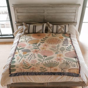 May include: A woven throw blanket with a floral pattern in shades of pink, yellow, and green. The blanket has a fringe border and is displayed on a bed with pillows.