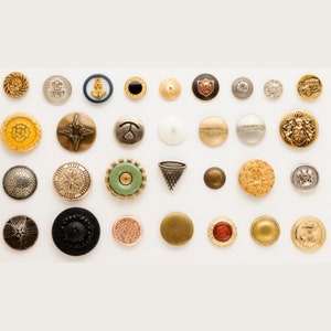 May include: An assortment of colourful and metallic buttons with various designs, including anchors, stars, and geometric shapes. The buttons are arranged in rows on a white background.