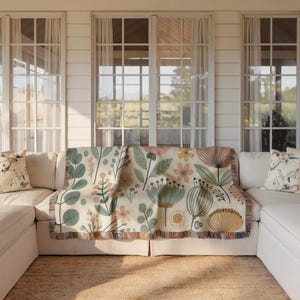 May include: A white couch with a floral patterned throw blanket. The throw blanket features a variety of flowers and leaves in shades of pink, green, and yellow. The couch is in a room with large windows and white walls.