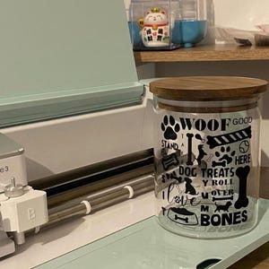Peut inclure: Un bocal en verre transparent avec un couvercle en bois, décoré de graphiques et de texte noirs sur le thème des chiens. Le bocal indique "Woof Good", "Dog Treats" et "Bones". Le bocal est posé sur une surface bleu clair.