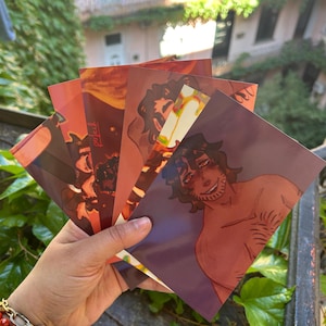May include: A hand holding a fan of art prints. The prints feature illustrations in shades of red, orange, and purple. The art style appears to be a mix of digital and traditional media. The prints are held in front of a background of greenery and buildings.