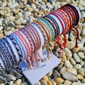 May include: A collection of colourful friendship bracelets made with woven string. The bracelets are displayed on a white stand and feature a variety of patterns and colours, including red, blue, green, yellow, pink, and white.