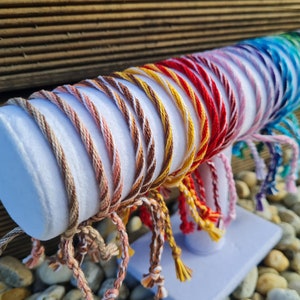 Op de afbeelding: Een selectie van kleurrijke vriendschapsarmbanden gemaakt van gevlochten touw, tentoongesteld op een witte standaard. De armbanden zijn gerangschikt in een regenboogpatroon, met kleuren die variëren van bruin tot blauw.