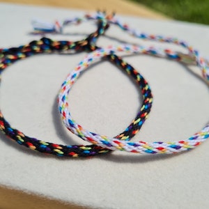 Pode incluir: Duas pulseiras de amizade, uma preta com detalhes arco-íris e outra branca com detalhes arco-íris.