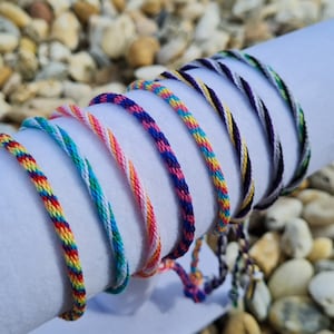 Op de afbeelding: Een set van acht kleurrijke vriendschapsarmbanden, elk met een ander streepjespatroon. De armbanden zijn gemaakt van dunne, gevlochten koorden in tinten rood, oranje, geel, groen, blauw, paars, wit en zwart.