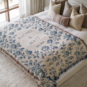 May include: A cream-coloured woven throw blanket with a blue floral pattern and the text "Rachel & Matt Est. 2023" in the centre. The blanket is draped over a white bed with pillows.