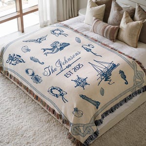 May include: A cream-coloured woven throw blanket with a nautical theme. The blanket features a mermaid, a sailboat, a lighthouse, a compass, a starfish, and a seashell. The text "The Johnsons EST 2025" is woven into the blanket.
