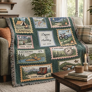 May include: A green throw blanket with a postage stamp design featuring outdoor activities like camping, hiking, and kayaking. The blanket includes the names "Logan & Ashley" and the year "EST. 2026". The blanket is draped over a sofa.