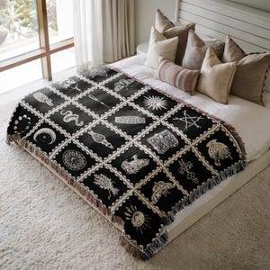 May include: Black and white woven throw blanket with a geometric pattern of various symbols, including moons, stars, and a snake. The blanket is fringed on all sides and is draped over a white bed.