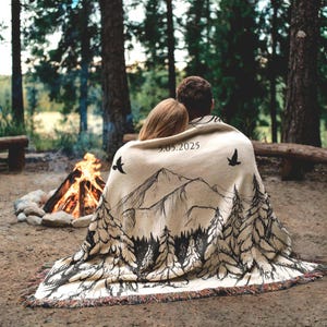 May include: A beige throw blanket with a black mountain and forest design, and the date 5.05.2025 printed on it. Two birds are also printed on the blanket. The blanket is draped over two people sitting near a campfire.