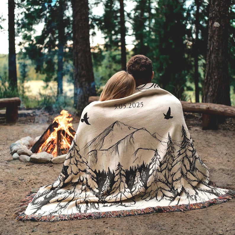 May include: A beige throw blanket with a black mountain and forest design, and the date 5.05.2025 printed on it. Two birds are also printed on the blanket. The blanket is draped over two people sitting near a campfire.