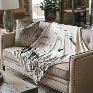 May include: A cream-colored blanket draped over a beige sofa. The blanket features a black and white mountain and forest design, with the names "Cery + Diane" and the date "5.05.2025" printed on it. A decorative pillow rests on the sofa.