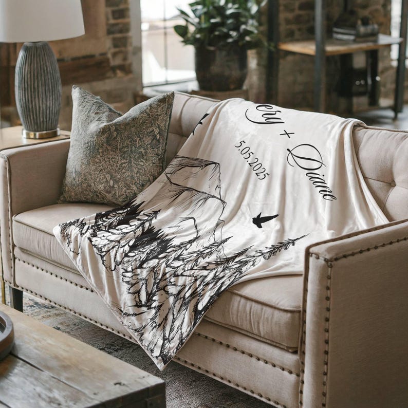 May include: A cream-colored blanket draped over a beige sofa. The blanket features a black and white mountain and forest design, with the names "Cery + Diane" and the date "5.05.2025" printed on it. A decorative pillow rests on the sofa.