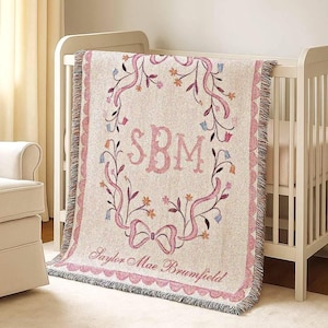 May include: A personalized baby blanket in a crib. The blanket is beige with a pink floral design and a pink border. It features the initials "SBM" in pink, surrounded by a floral wreath and the name "Saylor Mae Brumfield".