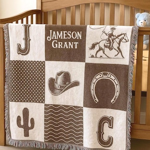 May include: A brown and white patchwork baby blanket with a cowboy theme. The blanket features a cowboy on a horse, a cowboy hat, a horseshoe, a cactus, and the name "Jameson Grant". The blanket is fringed.