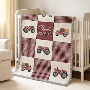 May include: A personalized baby blanket with a red and white checkered pattern. The blanket features red tractors and the name "Charlie JORDAN" in a script font. The blanket is draped over a white crib.