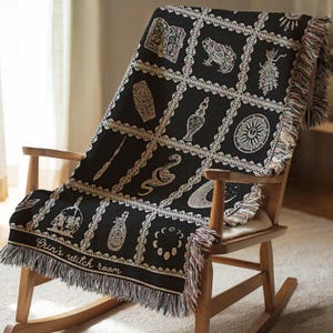 May include: Black woven throw blanket with a white design of various witchy symbols, including a crescent moon, a snake, a frog, a cauldron, and a bottle. The blanket has fringe on all sides and the text "Grin's witch room" is woven into the bottom corner.