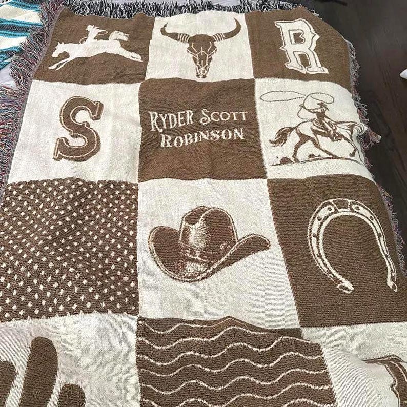 May include: A woven throw blanket featuring a grid of brown and cream panels with western-themed graphics including a cowboy on a horse, a bull skull, a horseshoe, a cowboy hat, the letter S, the letter R, and the text Ryder Scott Robinson.