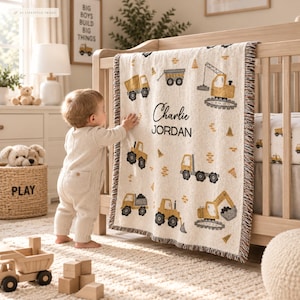Op de afbeelding: Een crèmekleurige geweven deken met een herhalend patroon van gele bouwvoertuigen, waaronder kiepwagens en graafmachines, met de gepersonaliseerde tekst Charlie JORDAN in het zwart gedrukt in het midden van de deken.